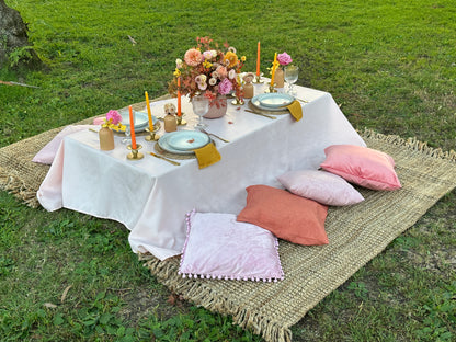 An outdoor picnic setup on a grassy lawn featuring a low table covered with a white tablecloth, set with plates, candles, and floral centerpieces, accompanied by several pastel throw pillows on a woven rug.