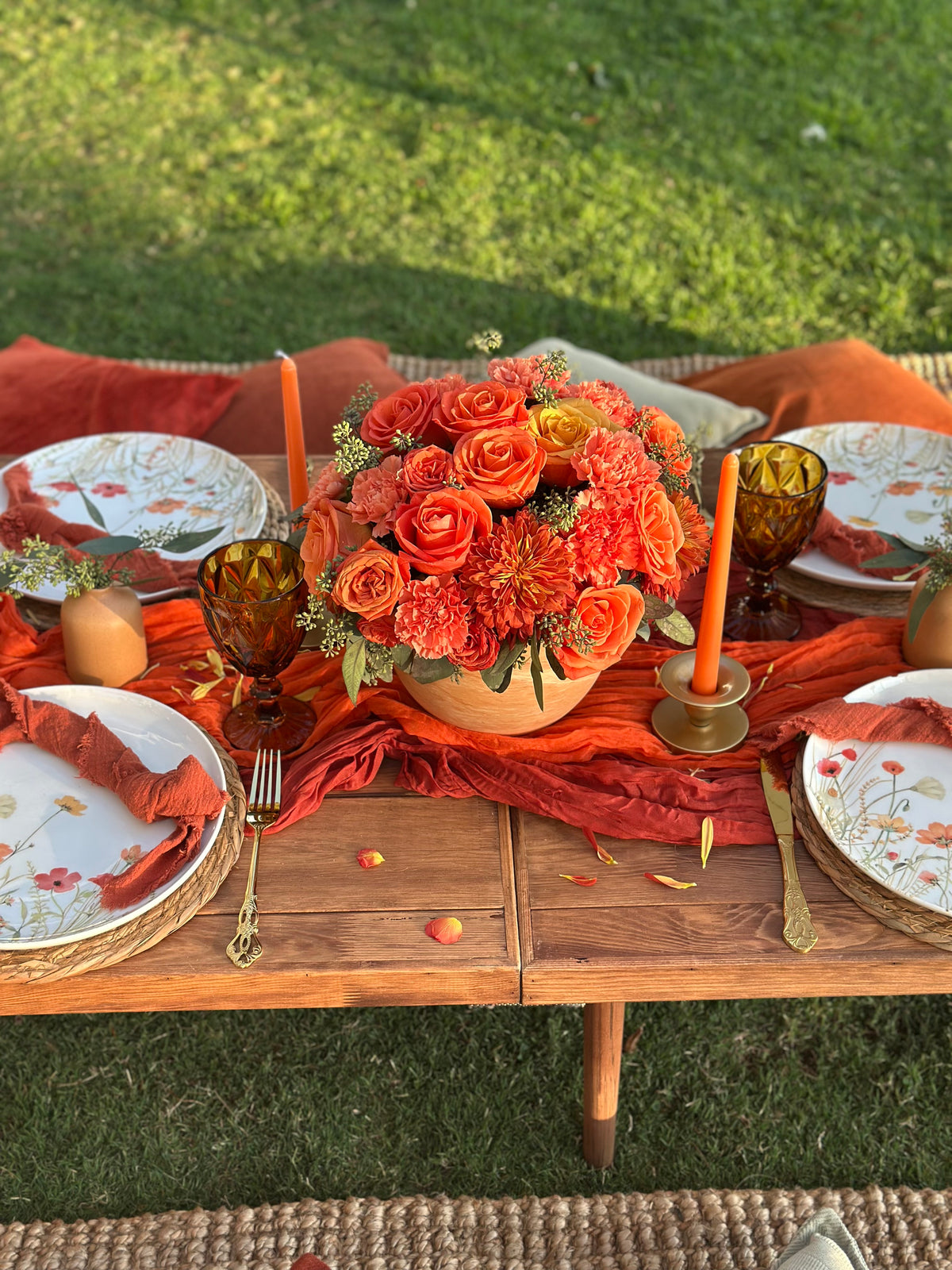 An outdoor autumn-themed table setting on a wooden table, featuring a large orange and red floral centerpiece, orange candles, patterned plates, and orange napkins on a green lawn background.