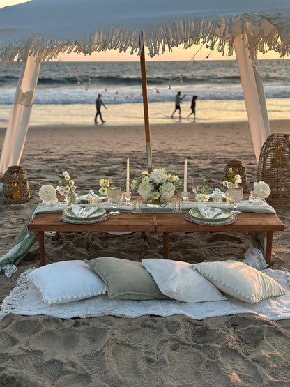 A beachside picnic setup on sand under a white canopy, with a low wooden table set for two, white flowers, candles, plates, and pillows. The ocean and distant beachgoers are visible in the background at sunset.