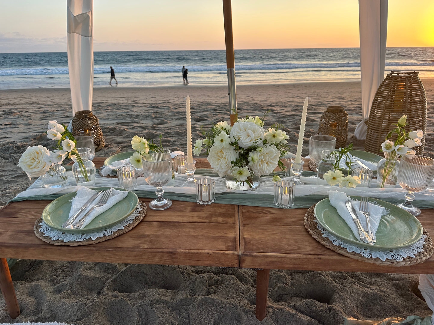 A beachside event table decorated with white floral arrangements, clear glassware, and light green plates under a wooden canopy with a rocky ceiling, overlooking the ocean at sunset.