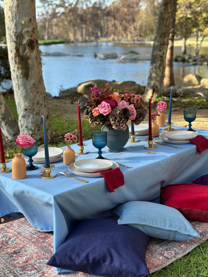 An outdoor table setting by a riverbank with a light blue tablecloth, white plates, blue and red glassware, a large floral centerpiece, and decorative cushions on the floor.
