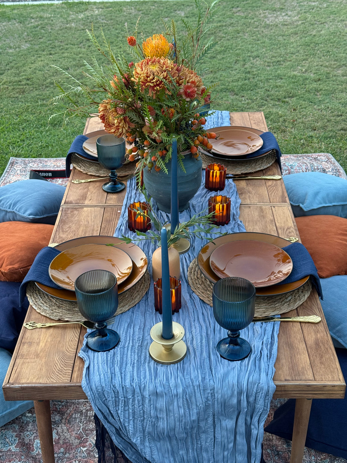 An outdoor dining table styled with a rustic wood table, a blue striped tablecloth, layered plates, dark blue goblets, amber glass candle holders, and a large autumnal floral centerpiece on a grassy lawn.