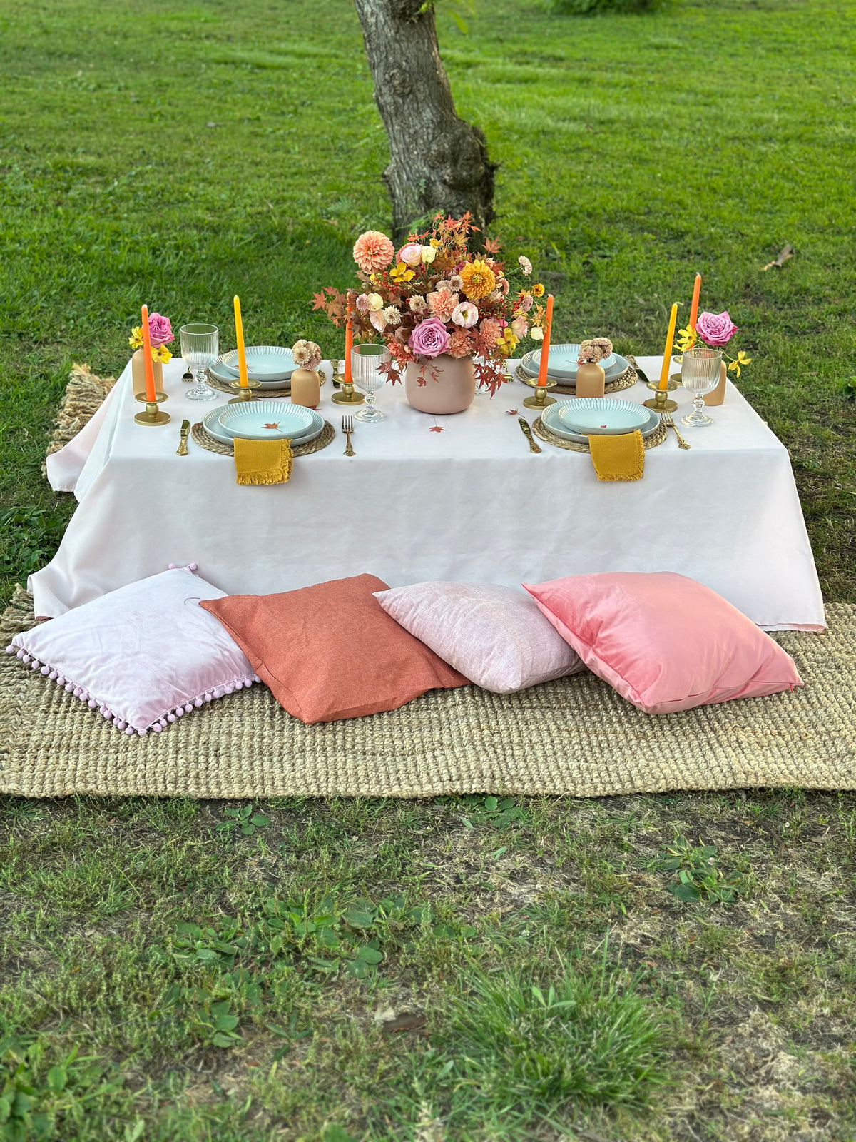 An outdoor picnic-style table setup on a grassy lawn under a tree, with a light pink tablecloth, floral centerpiece, candles, plates, and several pastel pillows arranged in front. 