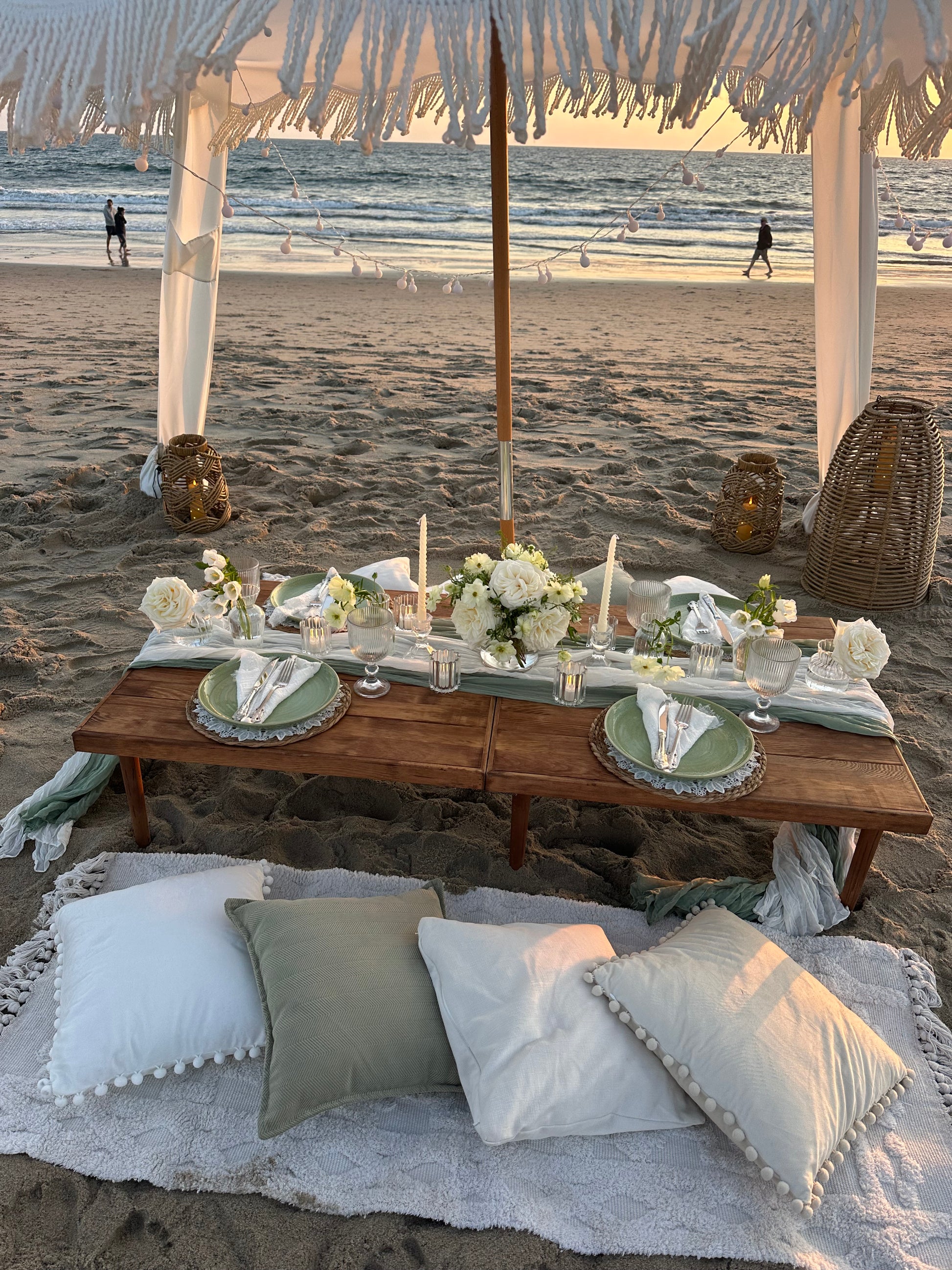 A beachside picnic setup under a white canopy on the sand, with a wooden table set for two, white flowers, candles, and plates. Comfortable white and sage green pillows are arranged on a white blanket in the foreground, with the ocean and distant beachgoers visible in the background.
