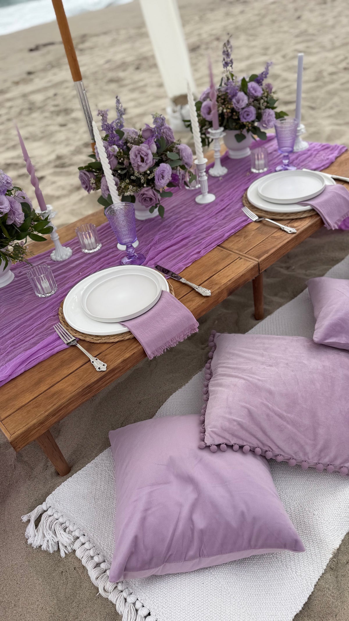 A beachside table setting styled in lavender purple, featuring a purple table runner, white plates, purple napkins, floral centerpieces, and matching purple pillows on a white fringed blanket on the sand.
