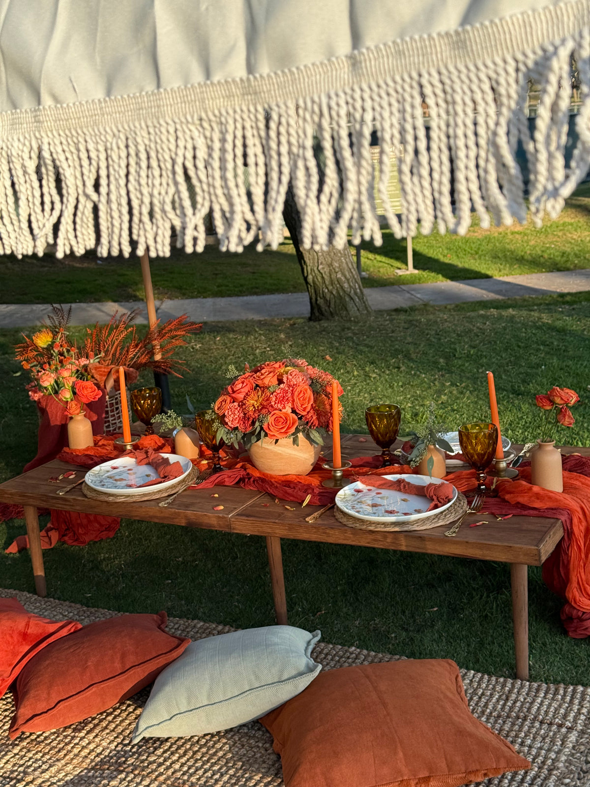 An outdoor table setting styled in autumn colors, featuring orange floral arrangements, candles, patterned plates, and rustic woven textiles on a grassy lawn.