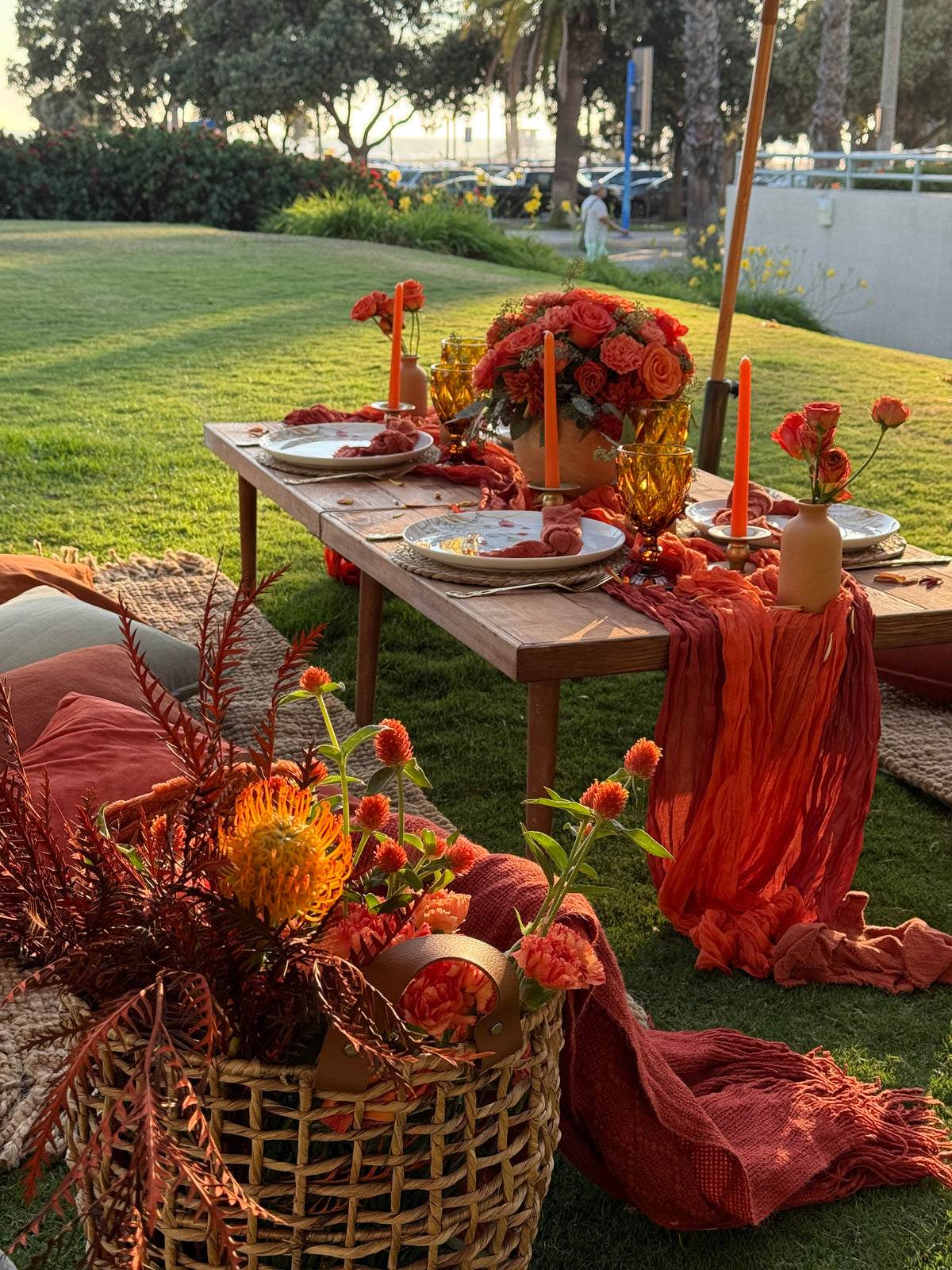 An outdoor autumn-themed table setting on a grassy lawn, decorated with rust-orange fabric drapes, orange and red flowers, candles, and a wicker basket with dried botanicals.