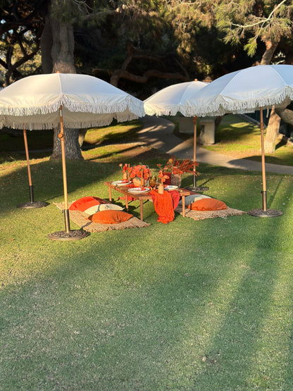 Outdoor park setup on a grassy lawn featuring two large white fringed umbrellas, a small table with orange textiles and floral arrangements, and several orange cushions on the grass.