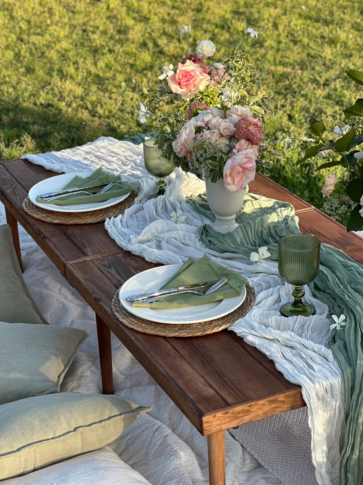 An outdoor dining table set with white plates, green napkins, woven placemats, a floral centerpiece, and draped white and sage green table runner on a grassy lawn.