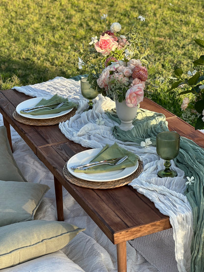An outdoor dining table set with white plates, green napkins, woven placemats, a floral centerpiece, and draped white and sage green table runner on a grassy lawn.