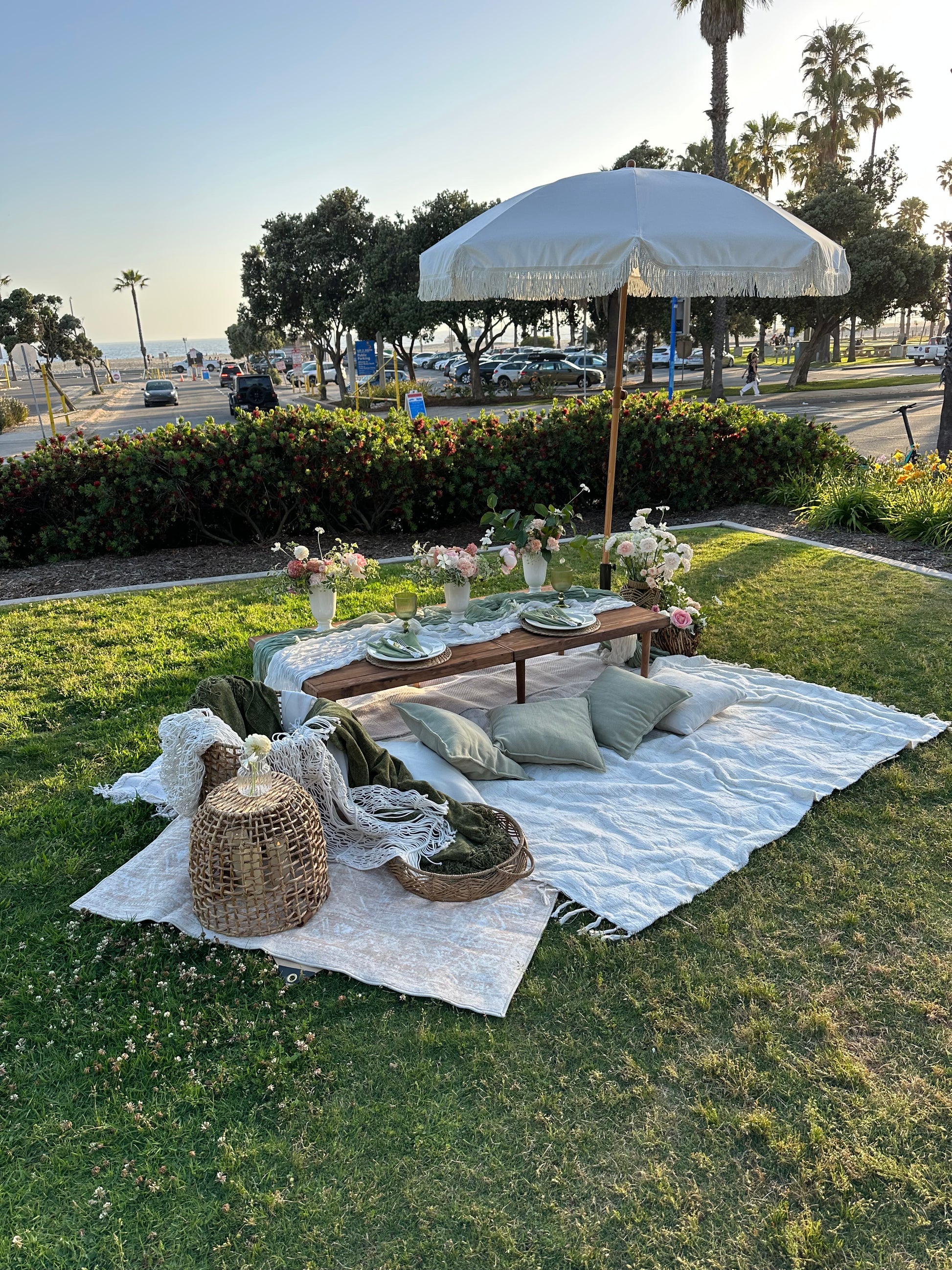 Outdoor picnic setup on a green lawn featuring a white picnic blanket, a wooden table with pillows, a white parasol, and woven baskets.