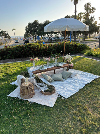 Outdoor picnic setup on a green lawn featuring a white picnic blanket, a wooden table with pillows, a white parasol, and woven baskets.