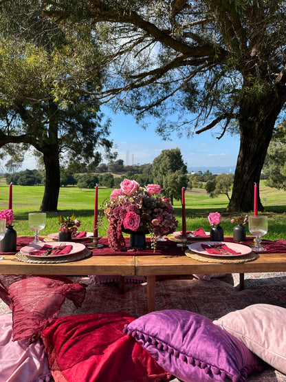 An outdoor dining setup with a wooden table set for four, featuring pink floral centerpieces, red candles, plates, and a lush green lawn and trees in the background.