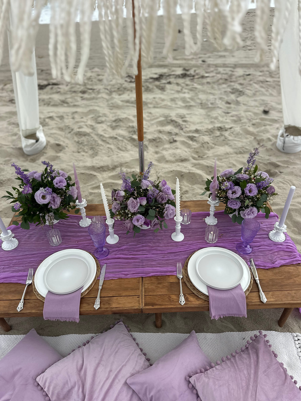 A styled beach table setup with a purple table runner, white plates, silver cutlery, purple flower arrangements, and white taper candles, with a wooden beach umbrella and white fringe canopy in the background.