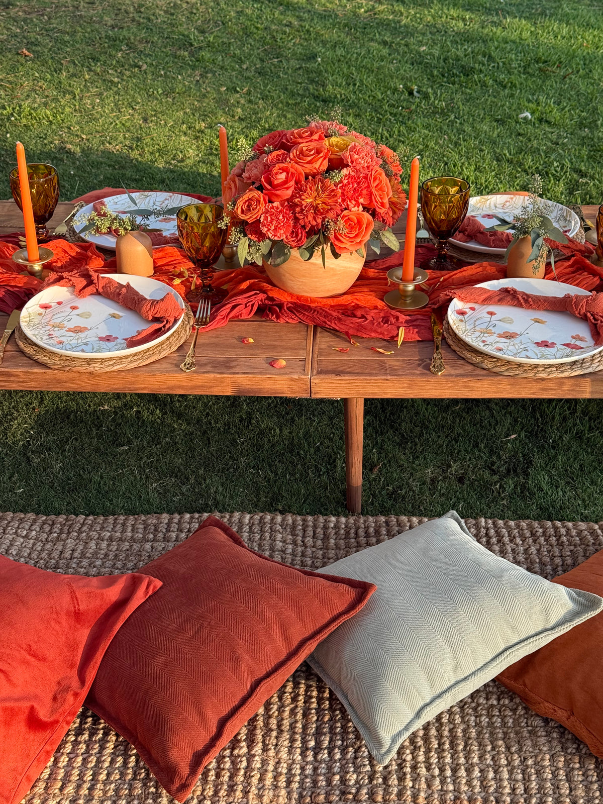 An outdoor autumn-themed table setting on a wooden table with orange floral centerpieces, orange plates, and orange candles, next to a woven bench with rust and cream pillows on a green lawn.