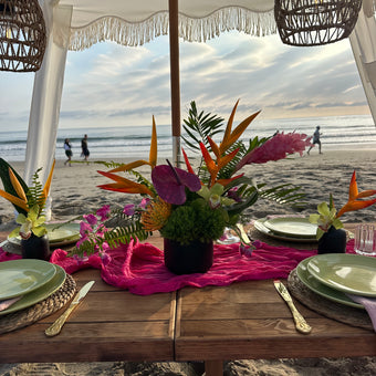 A beachside dining table decorated with vibrant tropical floral arrangements, a bright pink runner, green plates, and gold cutlery under a white canopy with wicker pendant lights.
