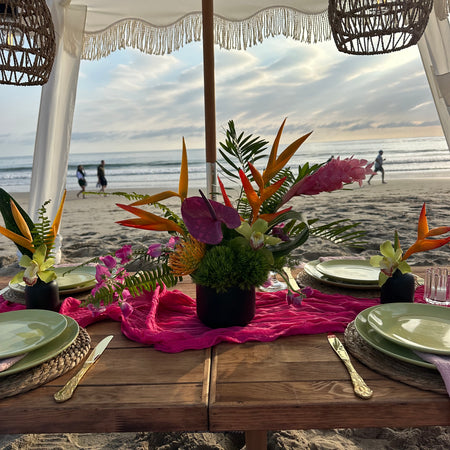 A beachside dining table decorated with vibrant tropical floral arrangements, a bright pink runner, green plates, and gold cutlery under a white canopy with wicker pendant lights.