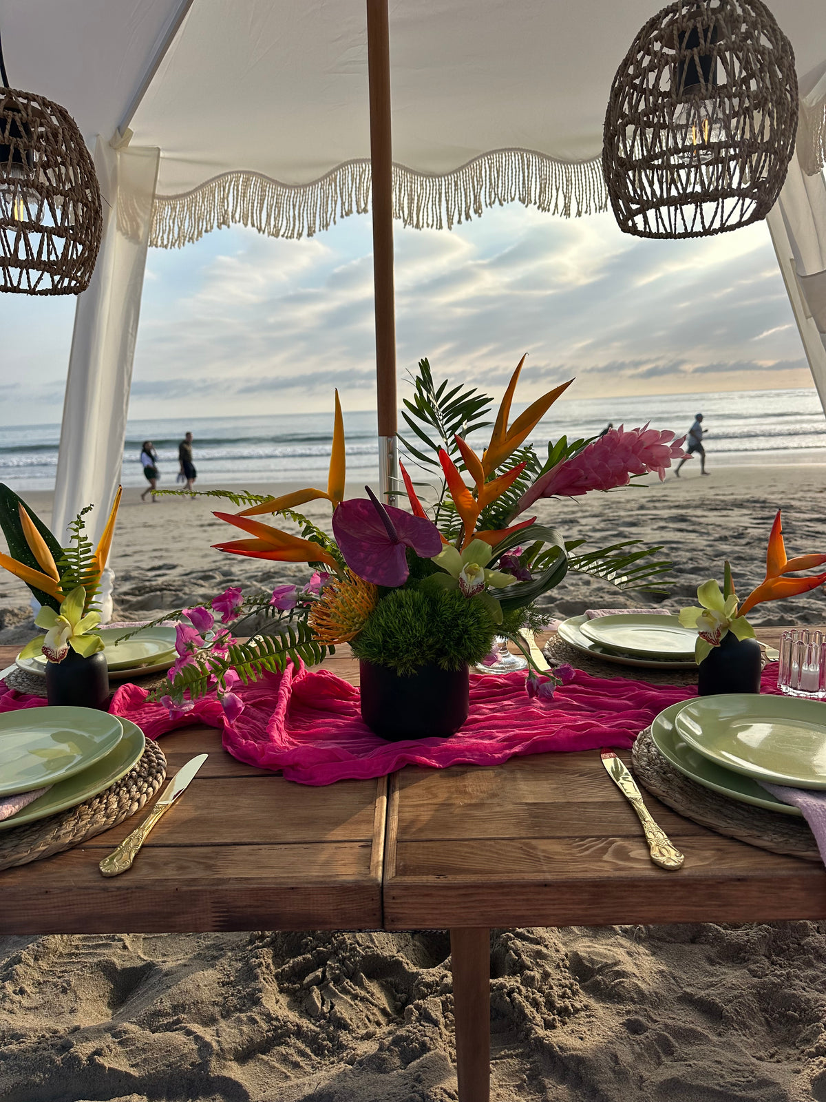 A beachside dining table decorated with vibrant tropical floral arrangements, a bright pink runner, green plates, and gold cutlery under a white canopy with wicker pendant lights.