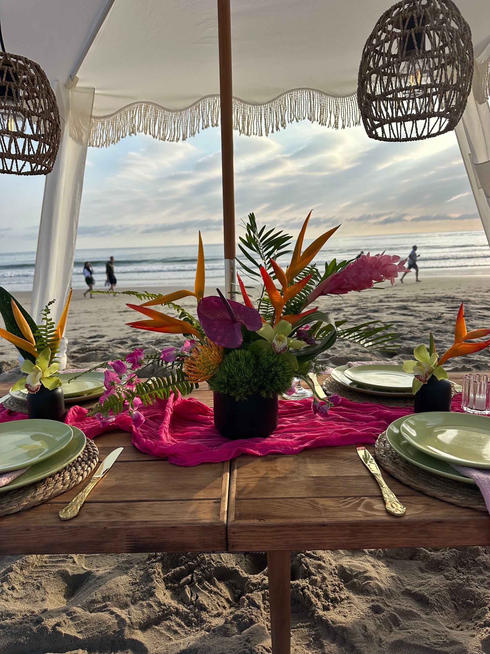 A beachside dining table decorated with vibrant tropical floral arrangements, a bright pink runner, green plates, and gold cutlery under a white canopy with wicker pendant lights.