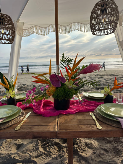 A beachside dining table decorated with vibrant tropical floral arrangements, a bright pink runner, green plates, and gold cutlery under a white canopy with wicker pendant lights.