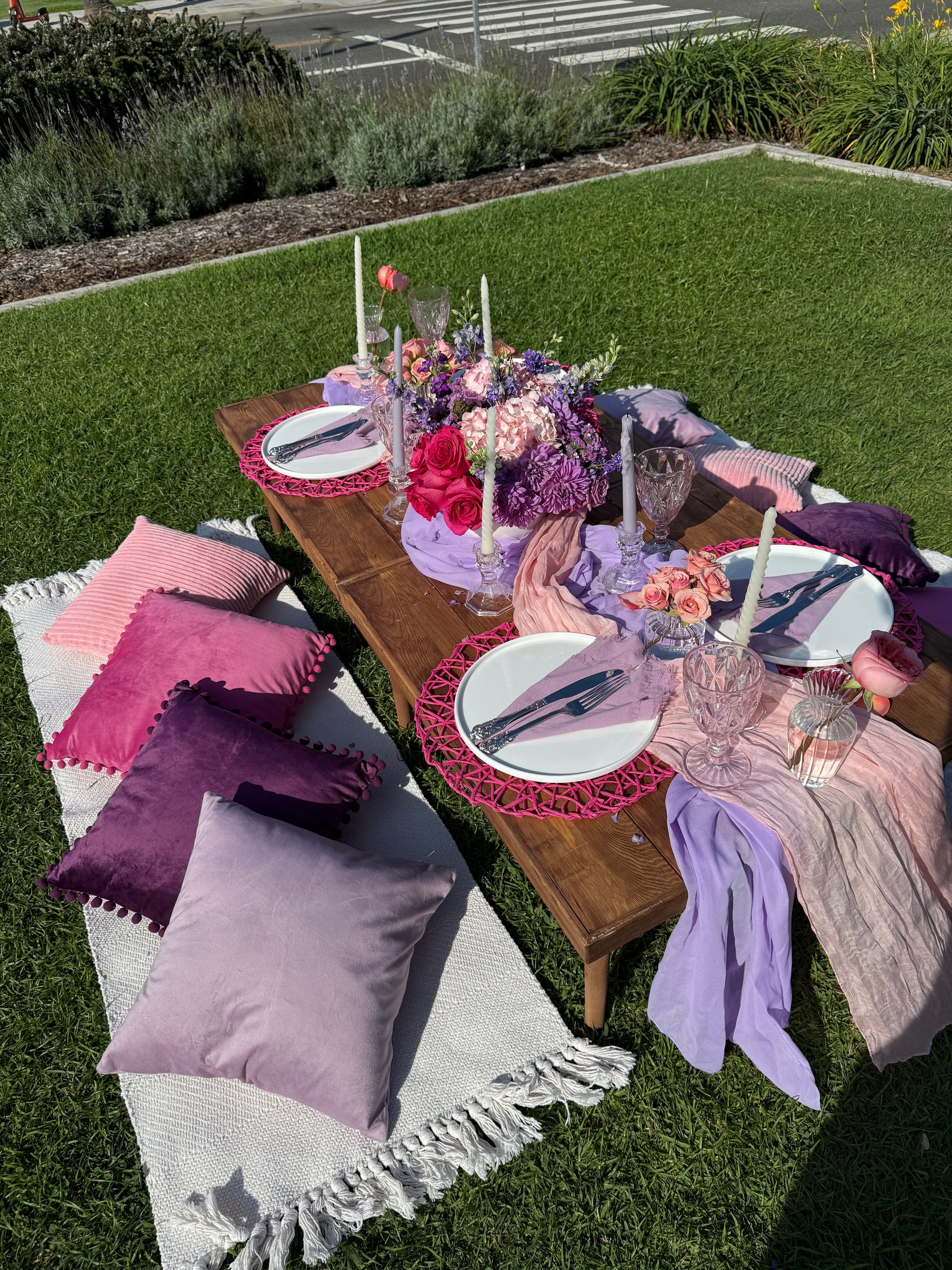 An outdoor dining setup on a wooden table with purple and pink table linens, white plates, floral centerpieces, and decorative pillows on a white fringed blanket on a green lawn.